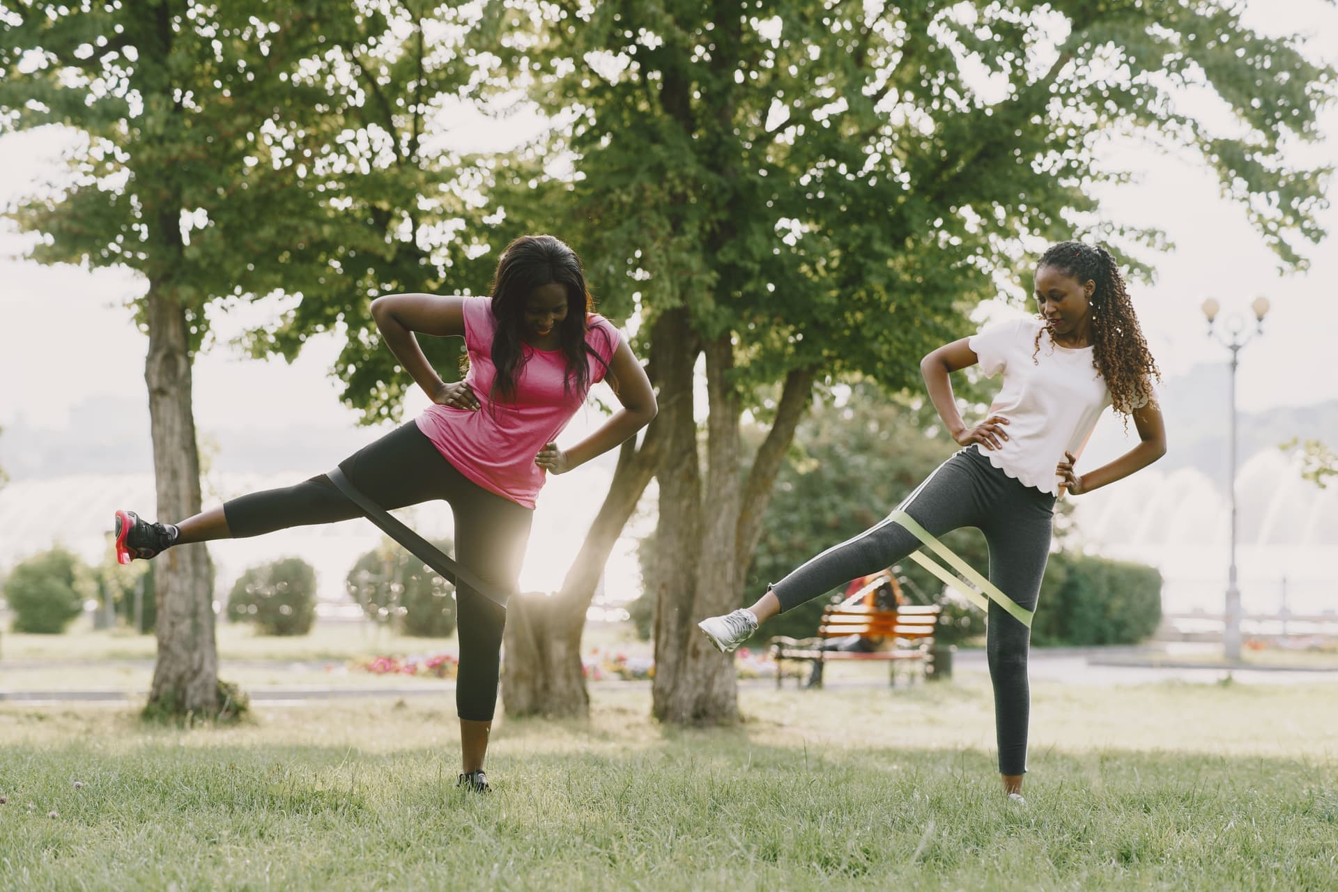 Two women doing banded leg raises together on the grass at a sunny park